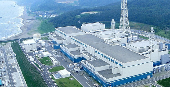 海岸沿いに建つ柏崎刈羽原子力発電所の空撮写真。白と青の建物群、送電塔、背景に緑の山々が見える